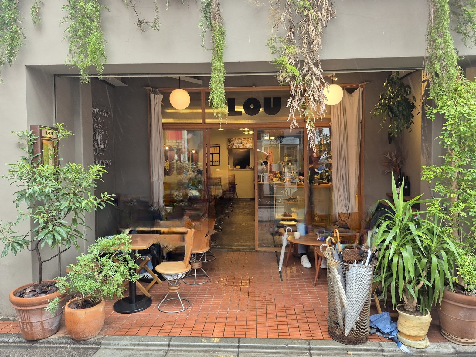 緑の鉢植えに囲まれたLOUの外観、ガラス越しに店内が見え、テラス席のテーブルと椅子が手前に並ぶ