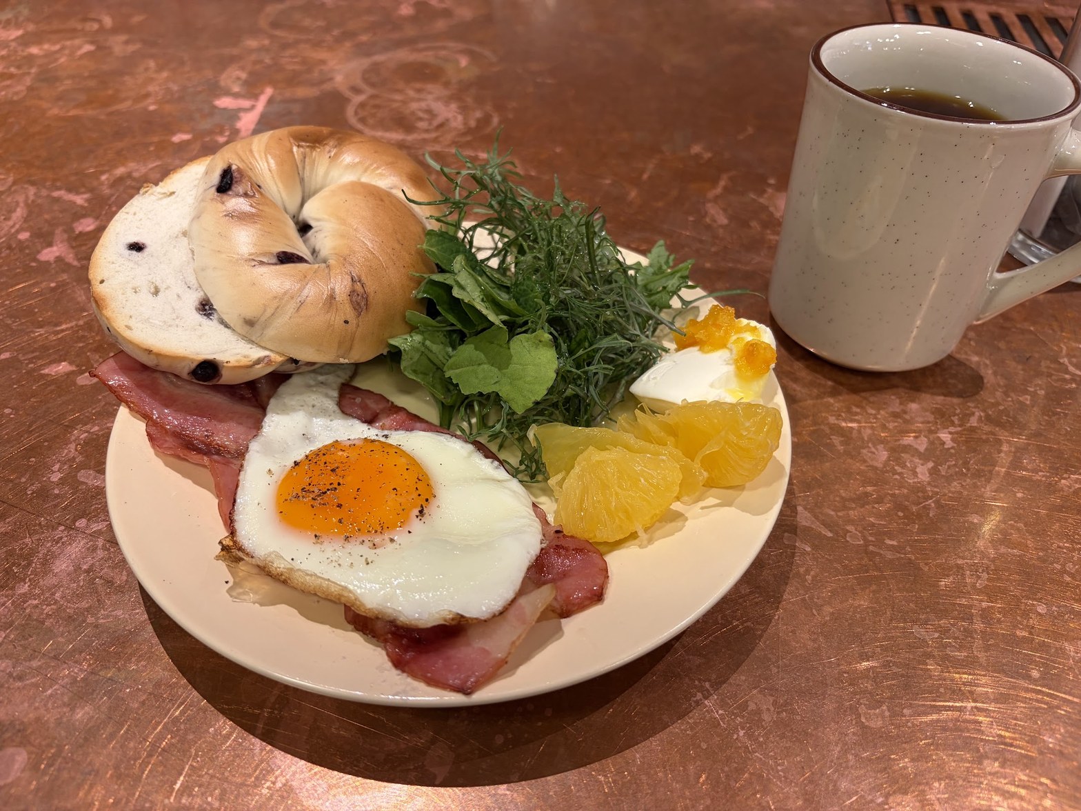 ベーグル・目玉焼き・ベーコン・グリーンサラダ・オレンジが盛られたプレートと白いマグカップ、ピンク大理石調カウンター上