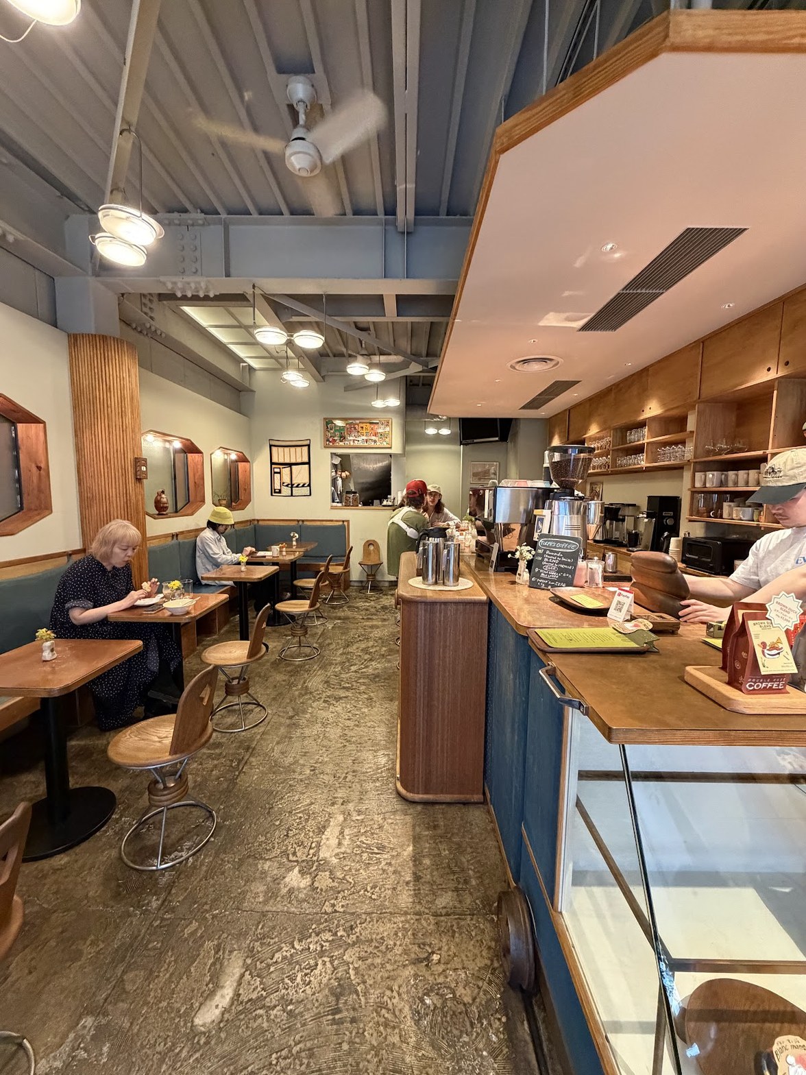 コンクリート床と木製テーブル・チェアが並ぶ店内、右手にカウンターとスタッフ、壁に絵画と鏡が点在