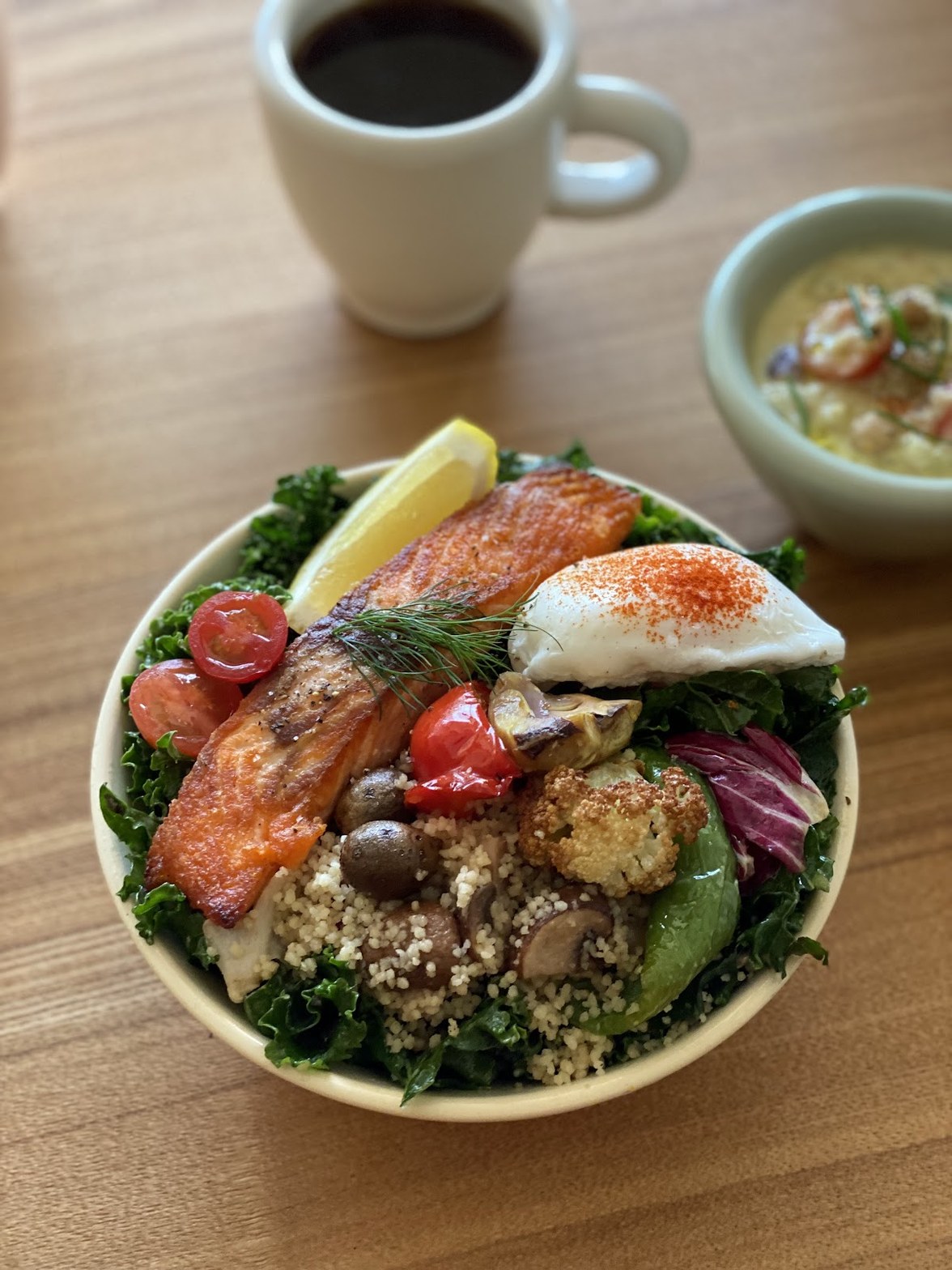 ケールと焼き鮭・ポーチドエッグ・ロースト野菜・クスクスが盛られたサラダボウル、奥に白いマグカップのブラックコーヒーと小鉢
